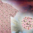 Load image into Gallery viewer, 3/4 Close Up view of the "Magnolia PI" 7-Strong Adult button down shirt, featuring white and green magnolia flowers patterned all over a light pink background. The shirt itself is displayed against a palm tree tropical background. The bottom left features a detail circle showing a close up of the shirt's floral design.