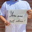 Load image into Gallery viewer, A midsection view of a male model wearing the 7-Strong "Stay" button down, a white shirt with staggered, hollowed out semicolons outlined in teal and purple - the colors for suicide awareness. Model is holding a handwritten sign saying "You Are Not Alone."