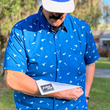 Load image into Gallery viewer, Male model wearing the adult 7-Strong "Paper Planes" button down shirt - featuring various sizes and designs of paper airplanes, some with dotted trails behind them against a royal blue background. Model is looking down admiring his paper airplane which displays the 7-Strong Brand logo.