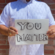 Load image into Gallery viewer, A midsection view of a male model wearing the 7-Strong "Stay" button down, a white shirt with staggered, hollowed out semicolons outlined in teal and purple - the colors for suicide awareness. Model is holding a handwritten sign saying "You Matter."