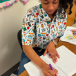 Load image into Gallery viewer, Female model sitting at a desk in a classroom, writing in a notebook, while wearing the 7-Strong Class Act shirt.