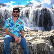 Load image into Gallery viewer, Male model, staged on the right of camera, sitting on rocks with a waterfall backdrop wearing the 7-Strong adult "Tropic of Conversation" button down - featuring various exotic rainforest flowers overlapping one another in a vibrant display against a white ghost background of other fronds and plants.