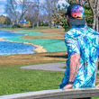 Load image into Gallery viewer, Male model, staged on the left of camera, staring out towards the water wearing the 7-Strong adult "Tropic of Conversation" button down - featuring various exotic rainforest flowers overlapping one another in a vibrant display against a white ghost background of other fronds and plants.