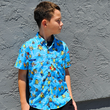 Load image into Gallery viewer, Youth male model standing against a gray stone wall, looking away from camera wearing the 7-Strong youth Avocado Coast button down.