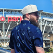 Load image into Gallery viewer, Male model in front of a baseball stadium wearing 7-Strong's adult Ballpark Blueprints shirt, featuring overhead maps of all the major league stadiums.
