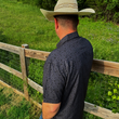 Load image into Gallery viewer, Male model wearing the 7-Strong Bandido adult short sleeve shirt featuring black and gray paisley design, along with a cowboy hat. Model has back facing the camer standing along a wood fence overlooking a field.