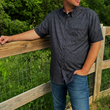 Load image into Gallery viewer, Male model, shot from the chin down, wearing the 7-Strong Bandido adult short sleeve shirt featuring black and gray paisley design. Model is looking away from camera, hand in pocket and standing along a wood fence overlooking a field.