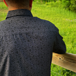 Load image into Gallery viewer, Male model wearing the 7-Strong Bandido adult short sleeve shirt featuring black and gray paisley design. Model has back facing the camera standing along a wood fence overlooking a field.