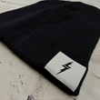 Load image into Gallery viewer, Angle full view of the black 7-Bolt beanie, rounded at the top and featuring a white 7-bolt tag in the bottom right corner of the cuff. The beanie is featured against a white brick backdrop.