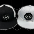 Load image into Gallery viewer, Both the black "Branded" trucker hat and the white "Branded" performance hat centered side-by-side against a black geometric background. Both feature raised black rubber X-Badge patches