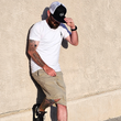 Load image into Gallery viewer, Male model, justified center of frame against a beige wall wearing the 7-Strong white "Branded" Tee and black 7-Strong "Branded" trucker hat, with raised X-Badge in black on the front. Model is looking down and away from camera.
