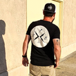 Load image into Gallery viewer, Male model with his back to the camera down an alley way, wearing the "Branded" black tee featuring the X-Badge logo centered in white on the back and wearing the "Branded" black trucker hat with the x-badge centered in black.