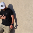 Load image into Gallery viewer, Male model, justified left of frame against a beige wall wearing the 7-Strong Black "Branded" Tee and white 7-Strong "Branded" white Performance hat, with raised X-Badge in black on the front. Model is looking down and away from camera.