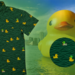 Load image into Gallery viewer, A 3/4 view of the 7-Strong "Duck O' The Irish" youth button down, featuring a deep green water-replicating background with various rubber ducks swimming along the waves. The shirt is featured against a green river scenic with a giant inflatable duck in the background. In the bottom right, there is a detail circle that shows the design pattern up close.