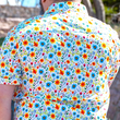 Load image into Gallery viewer, Behind the back look at a male model wearing the 7-Strong "Daisy Me Rollin'" adult button down shirt, a white base color with various blue, red, orange, and yellow floral daisies in an all over print. The model is facing away from camera standing out in nature.