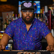 Load image into Gallery viewer, Male bartender standing behind the bar looking at the camera while wearing the 7-Strong "Happy Hour" button down shirt, featuring a multitude of neon colored drinks and glassware with garnishes, mixes, and the like all over a deep purple background.