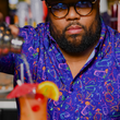 Load image into Gallery viewer, Male bartender crafting a mixed drink while wearing the 7-Strong "Happy Hour" button down shirt, featuring a multitude of neon colored drinks and glassware with garnishes, mixes, and the like all over a deep purple background.
