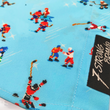 Load image into Gallery viewer, A bottom sweep tag view of the adult 8-Bit Hat Trick button down - featuring various 8-bit inspired scenes from hockey games on a light blue ice-like background. The shirt is featured against a ice hockey floor with hats strewn about.