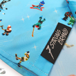 Load image into Gallery viewer, A bottom sweep tag view of the youth 8-Bit Hat Trick button down - featuring various 8-bit inspired scenes from hockey games on a light blue ice-like background. The shirt is featured against a ice hockey floor with hats strewn about.