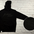 Load image into Gallery viewer, Full view of the 7-Strong Midnight Long Sleeve Hoodie, a black hoodie with black shadow font of the 7-Strong Brand logo. The hoodie is show against a white brick backdrop. The bottom right corner features a detail circle with the raised 7 Strong Brand logo across the chest.
