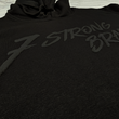 Load image into Gallery viewer, Midsection angled view of the 7-Strong Midnight Long Sleeve Hoodie, a black hoodie with black shadow font of the 7-Strong Brand logo. The hoodie is show against a white brick backdrop.