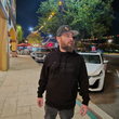 Load image into Gallery viewer, Male model out on the town looking off and wearing the 7-Strong Midnight Long Sleeve Hoodie, a black hoodie with black shadow font of the 7-Strong Brand logo.