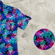 Load image into Gallery viewer, 3/4 close up view of the 7-Strong "Neon Coast" adult button down shirt. Featuring vibrant teal, pink, and blue palm fronds, overlapping throughout the shirt, against a black/deep purple background. The shirt is laid out on the sand of a beach. In the bottom right corner, there is a detail circle featuring a close up of the shirt's design.