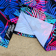 Load image into Gallery viewer, Bottom sweep tag view of the 7-Strong "Neon Coast" adult button down shirt. Featuring vibrant teal, pink, and blue palm fronds, overlapping throughout the shirt, against a black/deep purple background. The shirt is laid out on the sand of a beach.