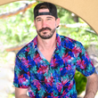Load image into Gallery viewer, Male model, facing camera with a slight smile, wearing the 7-Strong "Neon Coast" adult button down shirt. Featuring vibrant teal, pink, and blue palm fronds, overlapping throughout the shirt, against a black/deep purple background. Model is seated in an outside setting.
