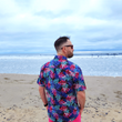 Load image into Gallery viewer, Male model, back to camera looking off in the distance on the beach, wearing the 7-Strong "Neon Coast" adult button down shirt. Featuring vibrant teal, pink, and blue palm fronds, overlapping throughout the shirt, against a black/deep purple background.