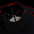 Load image into Gallery viewer, Centered underside look at the Black 7-Strong Sideline Script hat, with white 7-Bolts on the inside lining and black underbrim. Hat is on the back of a red and black fusion background.