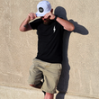 Load image into Gallery viewer, Male model, centered in frame against a beige wall wearing the 7-Strong Black "Branded" Tee and white 7-Strong "Branded" Performance hat, with raised X-Badge in black on the front. Model is looking down and away from camera.