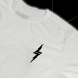 Load image into Gallery viewer, Front top of shoulder, left lapel look of the 7-Strong white Branded tee, featuring a 7-bolt left justified in black. Shirt is featured against a black geometric background.