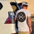 Load image into Gallery viewer, Male model against a car, back to camera wearing the white "Branded" X-Badge tee shirt, featuring a black X-badge logo centered on the back. on the left lapel - as well as wearing the "Branded" Trucker hat, with raised black X-Badge rubber patch
