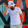 Load image into Gallery viewer, Male model against a colorful wall wearing the white "Branded" X-Badge tee shirt, featuring a black bolt on the left lapel - as well as wearing the white "Branded" performance hat, with raised black X-Badge rubber patch - model is looking down and away from camera, holding brim of hat with a hand in his pocket.