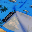 Load image into Gallery viewer, Bottom sweep tag view of the 7-Strong "7-Seas" shirt, a bright blue colored shirt with various nautical depictions such as islands, ships, mermaids, etc - drawn in a treasure map like fashion. The shirt is displayed against a weathered treasure map and compass background.