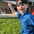Load image into Gallery viewer, A male child model wearing the youth 7-Strong "Paper Planes" button down shirt - featuring various sizes and designs of paper airplanes, some with dotted trails behind them against a royal blue background. The model is holding a paper airplane, smiling at camera, ready to throw the paper airplane.