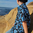 Load image into Gallery viewer, Side view of youth model sitting on rocks wearing the 7-Strong youth Electric Palms shirt, a collection of electric blue palm fronds featured against a black shirt. Model is overlooking the water.