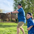 Load image into Gallery viewer, A father and son modeling of the adult and youth 7-Strong "Paper Planes" button down shirt - featuring various sizes and designs of paper airplanes, some with dotted trails behind them against a royal blue background. Both models are aiming to throw their paper planes.
