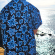 Load image into Gallery viewer, Back view of a youth model wearing the 7-Strong youth Electric Palms shirt, a collection of electric blue palm fronds featured against a black shirt. Model is displayed against an overlook of a scenic beach.