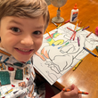 Load image into Gallery viewer, Male child model smiling into the camera at a table with school supplies and coloring pages, wearing the 7-Strong youth Class Act button down.