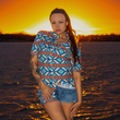 Load image into Gallery viewer, A person with long, dark hair stands by a body of water at sunset, facing at the camera. They are wearing a vibrant short-sleeve button-down shirt with an intricate Aztec-inspired pattern in blue, orange, and cream. The wind is blowing their hair as they look toward the horizon, where the sky glows with warm orange and pink hues