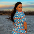 Load image into Gallery viewer, A person with long, dark hair and a confident expression stands by a body of water, turning slightly toward the camera. They are wearing a short-sleeve button-down shirt with an eye-catching Aztec-inspired pattern in blue, orange, and cream. The wind tousles their hair as the soft colors of a sunset cast a warm glow over the scene