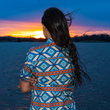Load image into Gallery viewer, "A person with long, dark hair stands by a body of water at sunset, facing away from the camera. They are wearing a vibrant short-sleeve button-down shirt with an intricate Aztec-inspired pattern in blue, orange, and cream. The wind is blowing their hair as they look toward the horizon, where the sky glows with warm orange and pink hues."