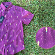 Load image into Gallery viewer, 3/4 Full view of the Cup of Life adult button up, a off-maroon colored shirt decorated throughout with falling gold confetti and depictions of the World Cup statue. The shirt is laid out on a grass pitch. In the bottom right corner is a detail circle, highlighting the shirt's design.