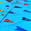Load image into Gallery viewer, Close up of midsection of the 7-Strong "Pennant to Win It" shirt, which is sky blue with texture, featuring various triangular flags featuring baseball's prominent cities patterned throughout. The shirt is featured against abackground is sunset at a major league stadium.