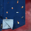 Load image into Gallery viewer, Bottom corner, sweep tag layout of "Call Me Old Fashioned" adult short sleeve button-down. Features elements of an Old Fashioned cocktail (bottle of bourbon, glasses, oranges, stirrers, and ice) against a blue background which faintly spells out an Old Fashioned recipe. Shirt is on a background featuring a finished Old Fashioned.