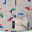 Load image into Gallery viewer, Close up of the bottom, sweep tag, portion of the Block Party shirt detailing the various colored blocks of different arrangements inspired by a classic video game, cascading down the shirt against a grayish background featuring shadows of the same shapes. The shirt is presented on a background of similar design.