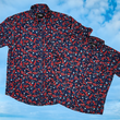 Load image into Gallery viewer, Full, overlapping view of the adult and youth 7-Strong "Aye Poppy" button down, featuring an array of red poppys with white sprigs on a deep navy blue shirt. The shirt is displayed against a partly cloudy sky.