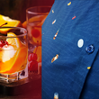 Load image into Gallery viewer, Side view layout of "Call Me Old Fashioned" adult short sleeve button-down. Features elements of an Old Fashioned cocktail (bottle of bourbon, glasses, oranges, stirrers, and ice) against a blue background which faintly spells out an Old Fashioned recipe. Shirt is on a background featuring a finished Old Fashioned and has a detail circle in the bottom corner showing design detail.